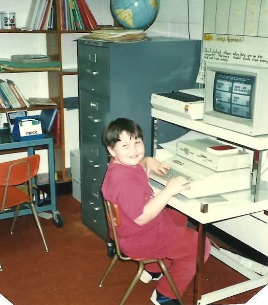 A young Jesse grinning at an Apple computer in a 1980s classroom
